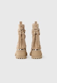 Bottines en daim beige avec des talons épais et des détails en franges accrochés à des anneaux en argent à l'arrière, sur un fond blanc.
