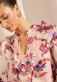 Gėlių blusa pasižymi šviesiai rožinės spalvos audiniu, raukšlėtomis briaunomis ir ryškiomis raudonomis, mėlynomis ir žalios spalvos raštais. V formos dekolte su surišimo detale.
