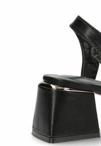 Sandalia de plataforma negra con un tacón grueso y geométrico y un acento metálico brillante en la base. Parte superior texturizada con una correa ajustable.