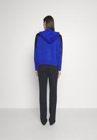 Kvinna som står vänd mot en vit vägg iförd en blå och svart huvtröja, svarta byxor och spetsiga klackar.