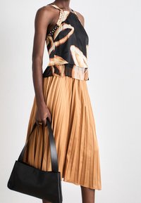 Top negro sin mangas con un patrón abstracto en beige, combinado con una falda plisada color beige. La modelo sostiene un bolso negro texturizado.