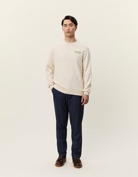 Beige pulóver zöld felirattal, kerek nyakkivágással és bordázott mandzsettával, sötétkék nadrággal és barna cipővel párosítva. Egyszerű háttér.