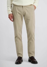 Man in beige chino broek, met bruine leren loafers en een crèmekleurige trui, staand met één hand in zijn zak tegen een effen achtergrond.