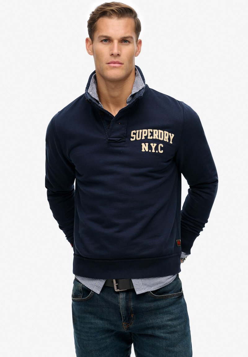 Sudadera marino con cuello parcial, logo "SUPERDRY N.Y.C." en beige, puños acanalados y una camisa a cuadros debajo. Jeans de mezclilla y cinturón negro.