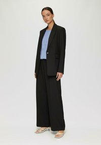Schwarzer maßgeschneiderter Anzug mit einem langen Blazer, weiter Hosen und einem hellblauen Oberteil. Glattes Material, minimalistisches Design und schlichte weiße High Heels.
