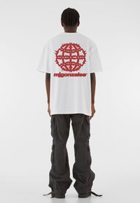 Valge ülerõhuga t-särk, mille seljal on suur punane graafika, mis sisaldab stiliseeritud kujundeid ja sõna "migonzfales", koos lõdva musta cargo-püksiga.