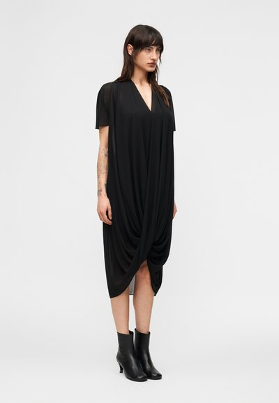 Γυναίκα που φοράει ένα χαλαρό μαύρο φόρεμα με κοντά μανίκια και ντεκολτέ σε σχήμα V, συνδυασμένο με μαύρα μποτάκια αστραγάλου, στέκεται μπροστά σε απλό λευκό φόντο.