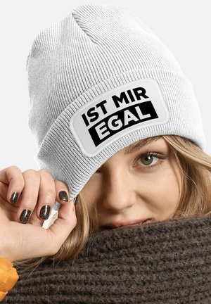 White knitted beanie with a ribbed texture, featuring a black patch that reads "IST MIR EGAL." The wearer holds a brown scarf.