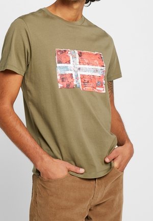 Homme portant un t-shirt vert olive avec un graphisme rouge, blanc et bleu délavé ressemblant à un drapeau norvégien usé. Mains dans les poches d’un pantalon marron.