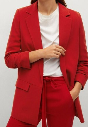 Kvinde iført en skrigrød blazer og matchende bukser med en hvid t-shirt med rund hals, hånd justerer blazer, neutralt baggrund.
