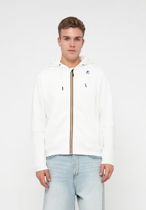 Young man wearing a white zip-up hoodie with multicolor zipper and light blue jeans, standing against a plain white background.