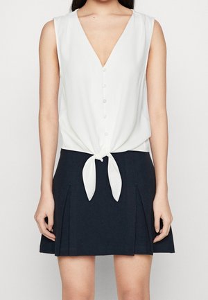 Haut blanc sans manches à boutons avec un nœud à l'avant, associé à une mini-jupe plissée bleu marine.