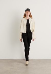 Korte crèmekleurige jas met frontzakken, zwart aansluitend shirt, zwarte slim jeans en beige schoenen, gecombineerd met een zwarte pet.