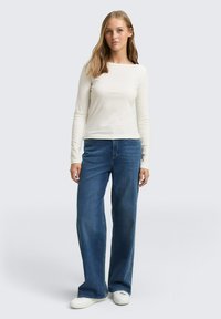 Witte ribbelige langemouwen top met een vierkante halslijn, gecombineerd met wijdvallende blauwe jeans en witte sneakers. Eenvoudige, casual outfit.