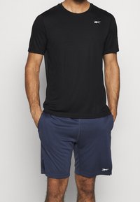 Camiseta atlética negra de manga corta con cuello redondo, combinada con pantalones cortos azul marino que tienen una cinturilla elástica y bolsillos laterales. Logotipo blanco en la camiseta.