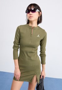 Olivinis žalias ilgas rankoves džinsinis suknelė su išilginiu strukturiniu raštu ir šonine plyšio, ant krūtinės esantis baltas Air Jordan logotipas.