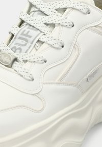 Sneaker bianca con tomaia in pelle liscia, suola spessa e lacci grigi. Presenta un design testurizzato e il logo "Buffalo" sulla linguetta.