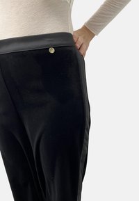 Sorte bukser i et glat stof, med en blank talje og et lille guld hardware-detalje. Designet inkluderer en sideliste.