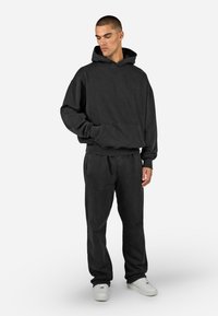 Čierna mikina s kapucňou a zodpovedajúce tepláky vyrobené z mäkkej textílie. Má prednú kapsu, rebrované manžety a voľný strih. Biele tenisky dopĺňajú outfit.