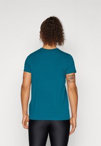 Person mit lockigem Haar, die ein türkisfarbenes kurzärmeliges Hemd und schwarze Leggings trägt, mit dem Rücken zur Kamera und sichtbaren Tattoos auf beiden Oberarmen.