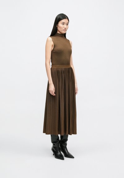 Καφέ ριχτό πλεκτό φόρεμα με ψηλό λαιμό και χωρίς μανίκια, που διαθέτει εφαρμοστό κορσέ και ρέουσα πλισέ φούστα. Μαύρες μακριές μπότες.