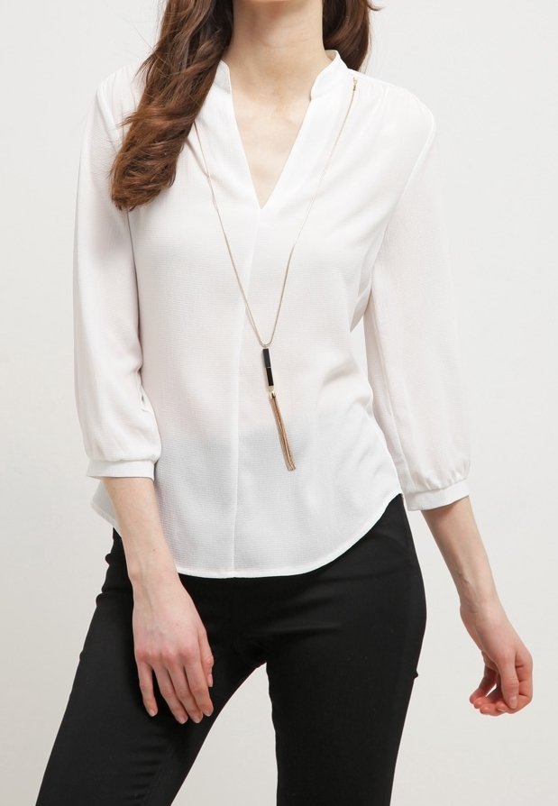 Femme portant un chemisier blanc à col en V avec des manches trois-quarts et un long pendentif, associé à un pantalon noir.