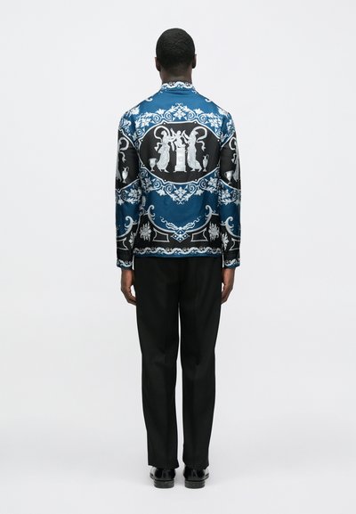 Modrá a čierna košeľa s dlhým rukávom s lesklým povrchom, zdobená prepracovanými bielymi vzormi, kombinovaná s prispôsobenými čiernymi nohavicami.