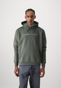 Zelená bavlnená mikina s kapucňou a prednou vreckom. Obsahuje biele logo "TOMMY JEANS" napísané na hrudi. Model má na sebe šedé džínsy.
