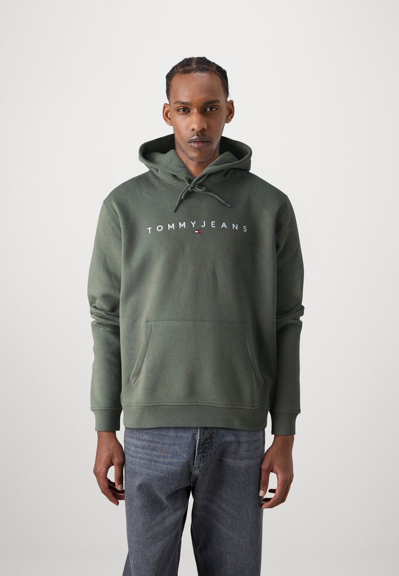 Žalias medvilnės hoodie su priekinėmis kišenėmis ir raišteliu ant gobtuvo. Ant krūtinės balta spalva užrašyta "TOMMY JEANS" logotipas. Modelis vilki pilkas džinsus.