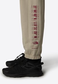 Pantalones de chándal beige con texto impreso en rojo oscuro y puños elásticos, combinados con zapatillas deportivas negras diseñadas para uso exterior.