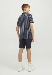 Camisa de manga corta para niños con rayas verticales en azul marino y blanco, combinada con pantalones cortos azul marino y zapatillas blancas, vista desde la parte de atrás.