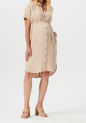 Robe chemise beige à manches courtes, cintrée à la taille, dotée d'une ceinture à nouer et d'un ourlet arrondi. Portée avec des sandales à talons carrés beiges.
