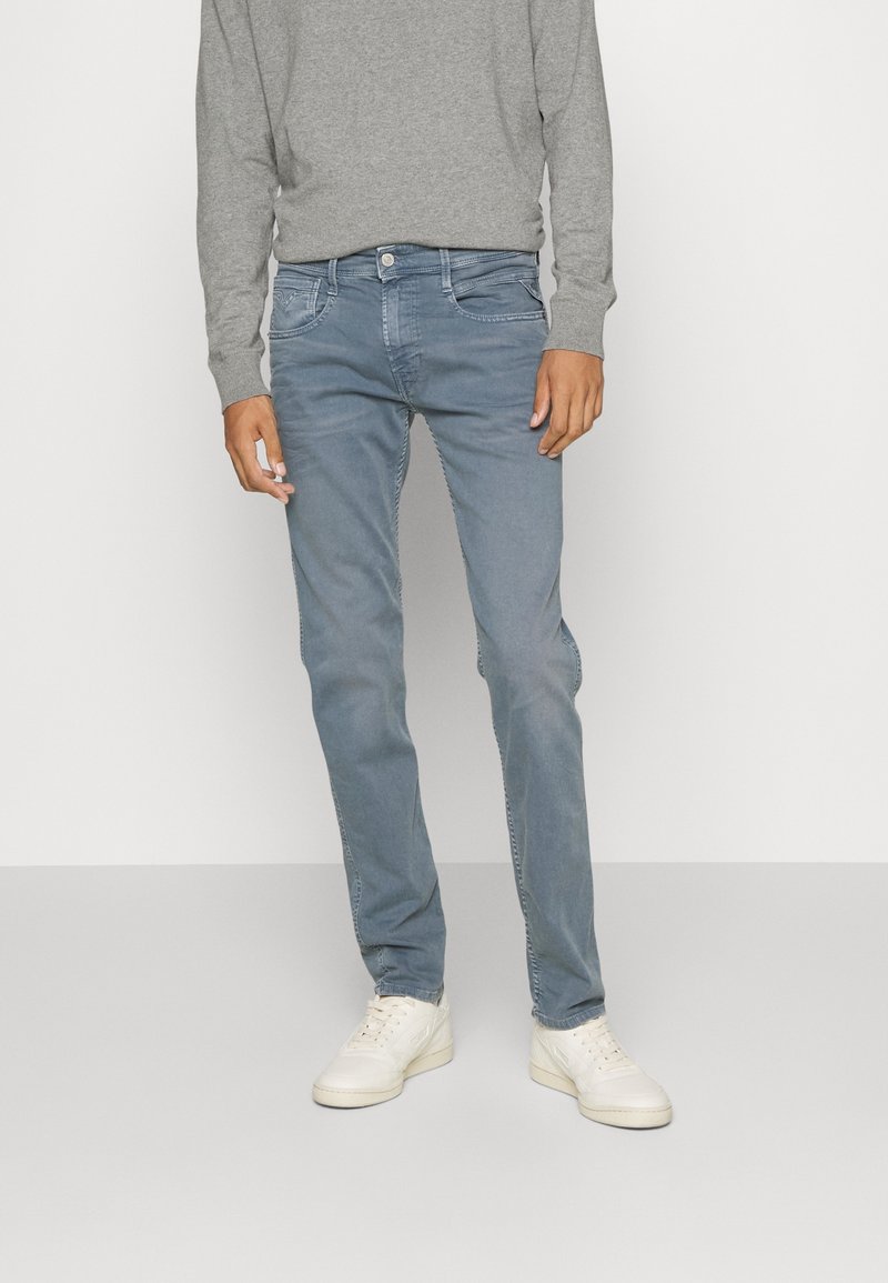 Homme portant un jean bleu délavé, un pull gris clair à manches longues et des baskets blanches, debout devant un fond uni.