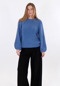 Žena stojaca v modrom hrubom pletenom svetri s balónovými rukávmi a čiernych širokých nohaviciach na hladkom pozadí.