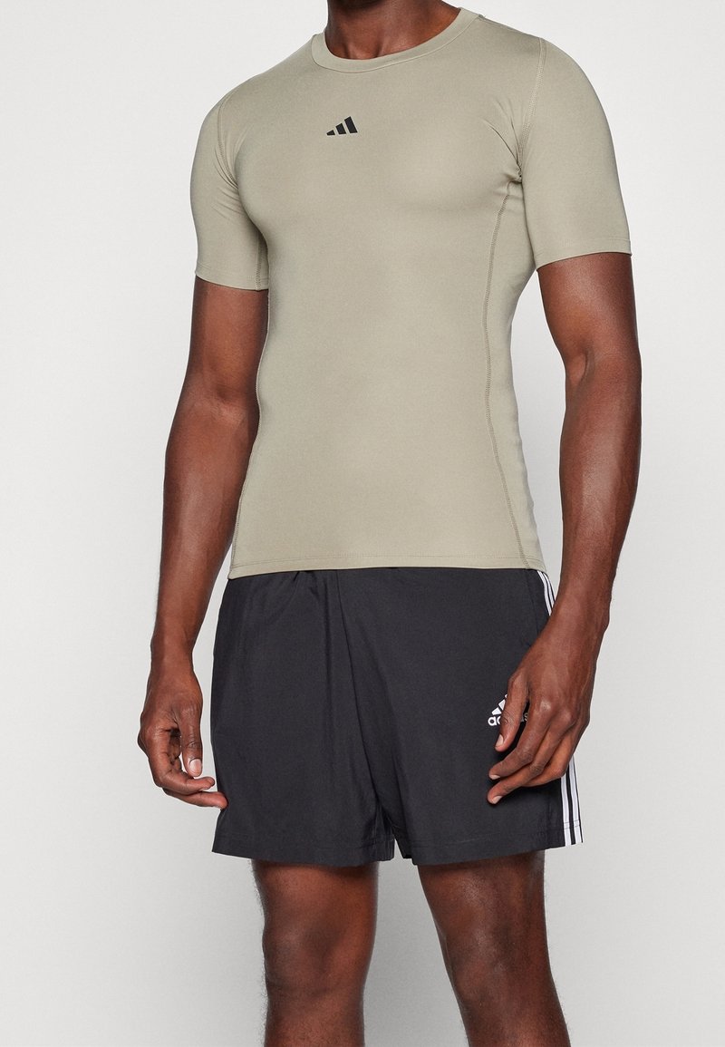 Beżowa koszulka sportowa z krótkim rękawem z czarnym logo na klatce piersiowej, połączona z czarnymi szortami z trzema białymi paskami i logo z boku.