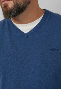 Blauwe v-hals trui van zacht breiwerk, met een klein logo op de linkerkant van de borst en gedragen over een witte ronde hals t-shirt.
