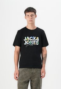 Black cotton t-shirt featuring multi-colored "JACK & JONES" text and graphic design. Model wearing olive green textured pants.