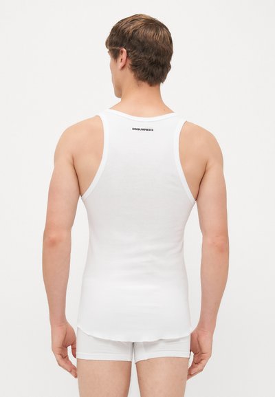 Mann steht mit dem Rücken zu uns und trägt ein weißes geripptes Tanktop mit dem "DSQUARED2"-Logo und passender weißer Boxershorts.