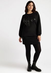 Sudadera negra con aplicaciones de corazones en capas, combinada con una falda negra y medias. El calzado incluye botas negras hasta el tobillo con suela rugosa.