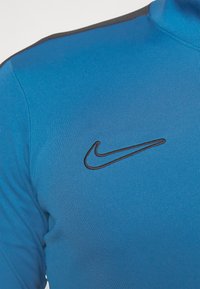 Nike plavi sportski majica s visokim ovratnikom, teksturiranom tkaninom i crnim logotipom na lijevoj strani prsnog koša. Naglasci u tamnijoj nijansi duž ramena.