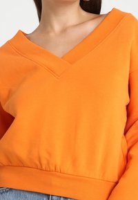 Pull-over orange avec un décolleté en V profond, une bordure côtelée et des manches longues ; fabriqué en tissu doux et texturé avec une coupe décontractée.