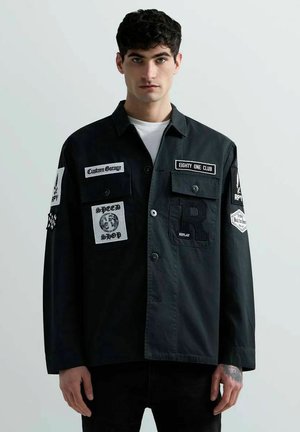 Giovane uomo con capelli ricci scuri indossa una giacca nera con numerose toppe e loghi bianchi su sfondo chiaro e uniforme.