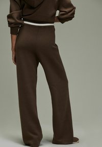 Brązowe, dziane spodnie o szerokich nogawkach z delikatną teksturą, z kontrastującym białym paskiem w talii. Krój jest luźny i swobodny.