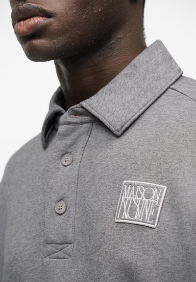 Κοντινό πλάνο ενός ατόμου που φοράει γκρι πόλο πουκάμισο με γιακά, δύο κουμπιά και ένα λευκό πατς "Maison Kitsuné" στο στήθος.