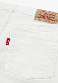 Valged puuvillased denim teksased, millel on punane silt tagataskus ja pruuni nahast etikett, millel on reljeefne graafika ja tekst.