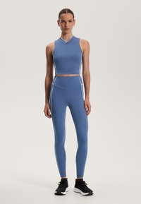 Modra športna športna majica s belim pipingom, kombinirana z visokim pasom hlačami. Obe sta izdelani iz raztegljivega materiala, zasnovana za aktivno nošenje. Črne superge.