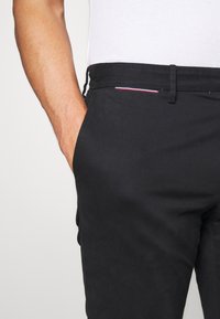 Svarta byxor i slätt tyg med rak midja, med en sidoficka och en liten detalj i röd, vit och blå kantstyg.