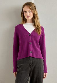 Purple ribbed cardigan with a V-neck and five-button closure, featuring long sleeves and a cropped design. Paired with black pants.