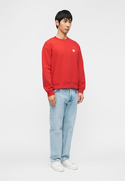 Νεαρός άνδρας που στέκεται φορώντας κόκκινο φούτερ με μικρό λευκό λογότυπο, ανοιχτόχρωμο μπλε τζιν και λευκά αθλητικά σε απλό γκρι φόντο.