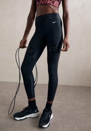 Atleta che indossa leggings neri Nike e sneakers, con in mano una corda per saltare, in piedi su un pavimento grigio davanti a una parete semplice.
